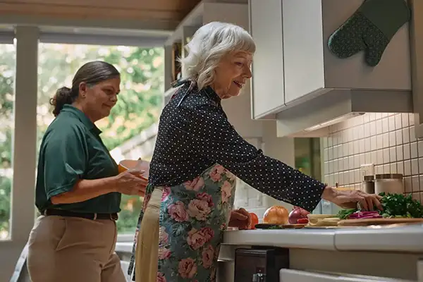A Home Instead caregiver and an elderly woman prepare a meal together in a sunny kitchen, symbolizing teamwork and independence in home care.